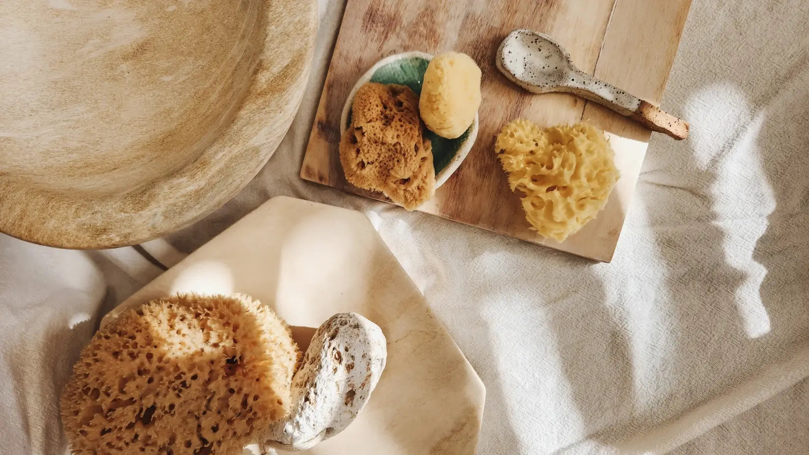 Natural sea sponges on a wooden cutting board with a wooden spoon and a ceramic dish on a light fabric background.